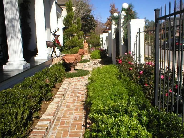 Brick walkway between boxwood hedges and roses beside a columned porch with a spiral topiary