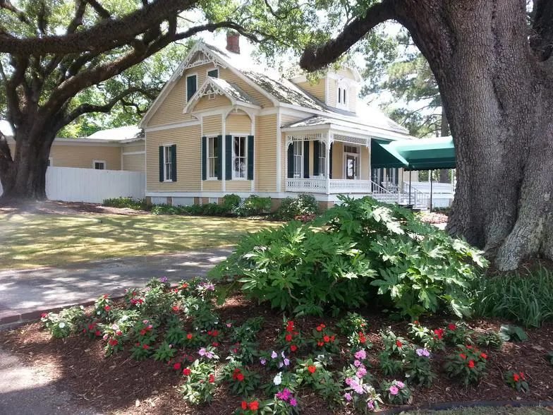Yellow Victorian house under live oaks with a bed of red and pink vinca and fatsia