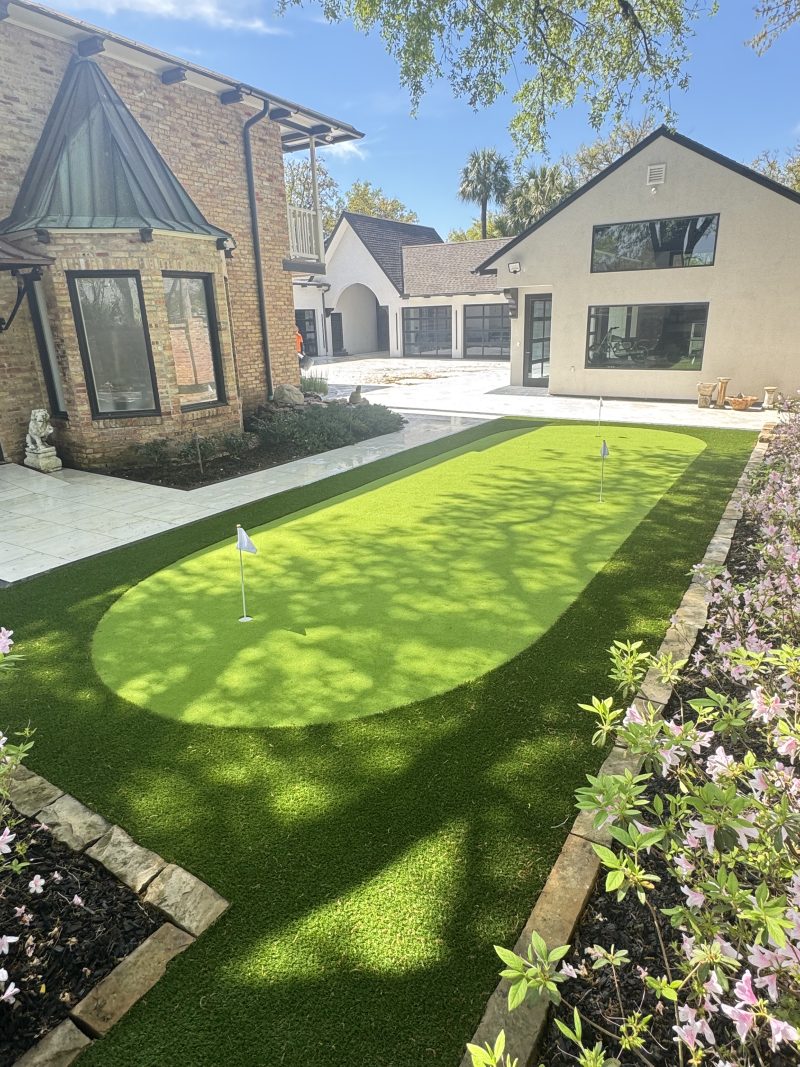 Artificial turf putting green with flags beside a brick home, edged with azaleas