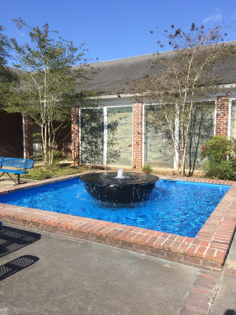 Square fountain pool with a black bowl fountain in a brick courtyard, river birches behind