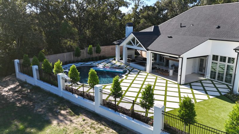 Aerial view of a pool deck with turf strips between pavers, a white columned fence and a row of Italian cypress