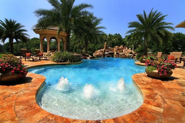 Freeform pool with bubblers, potted red flowers, palms and a stone gazebo beside a rock waterfall