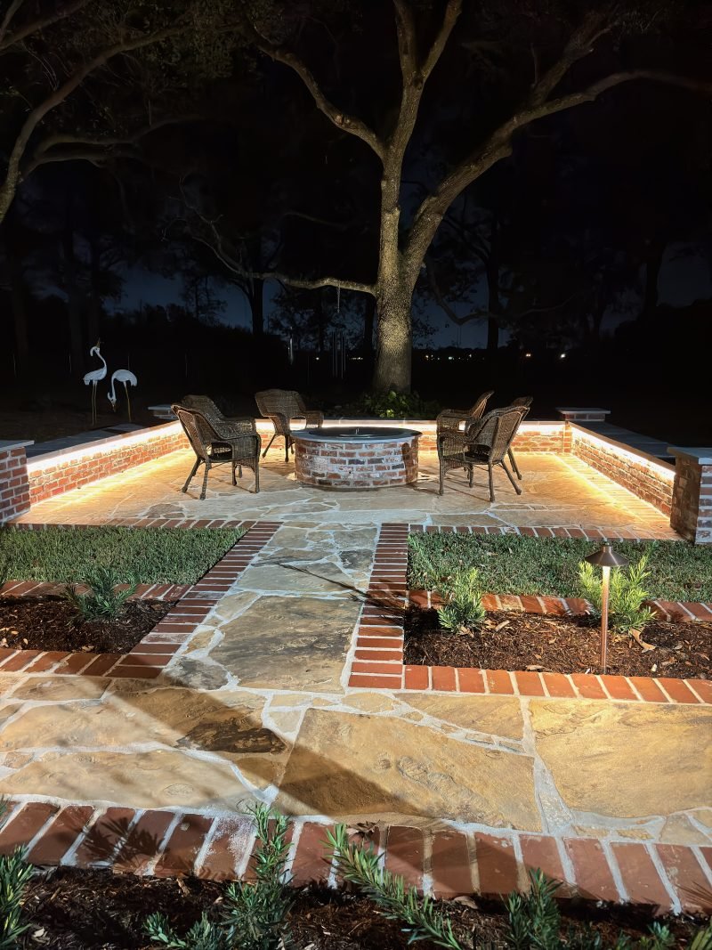 Flagstone patio at night with a lit fire pit seating area and brick-edged beds with path lights