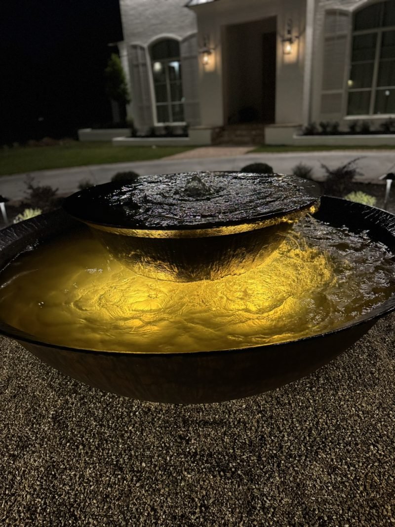 Fountain bowl lit from inside, spilling into a basin in front of a white home at night