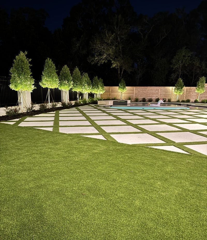 Lit trees, turf lawn and paver patio around a pool at night