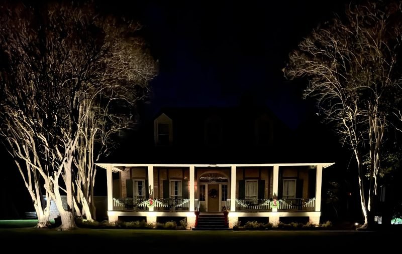 Front of a raised porch home at night with uplit crepe myrtles on each side